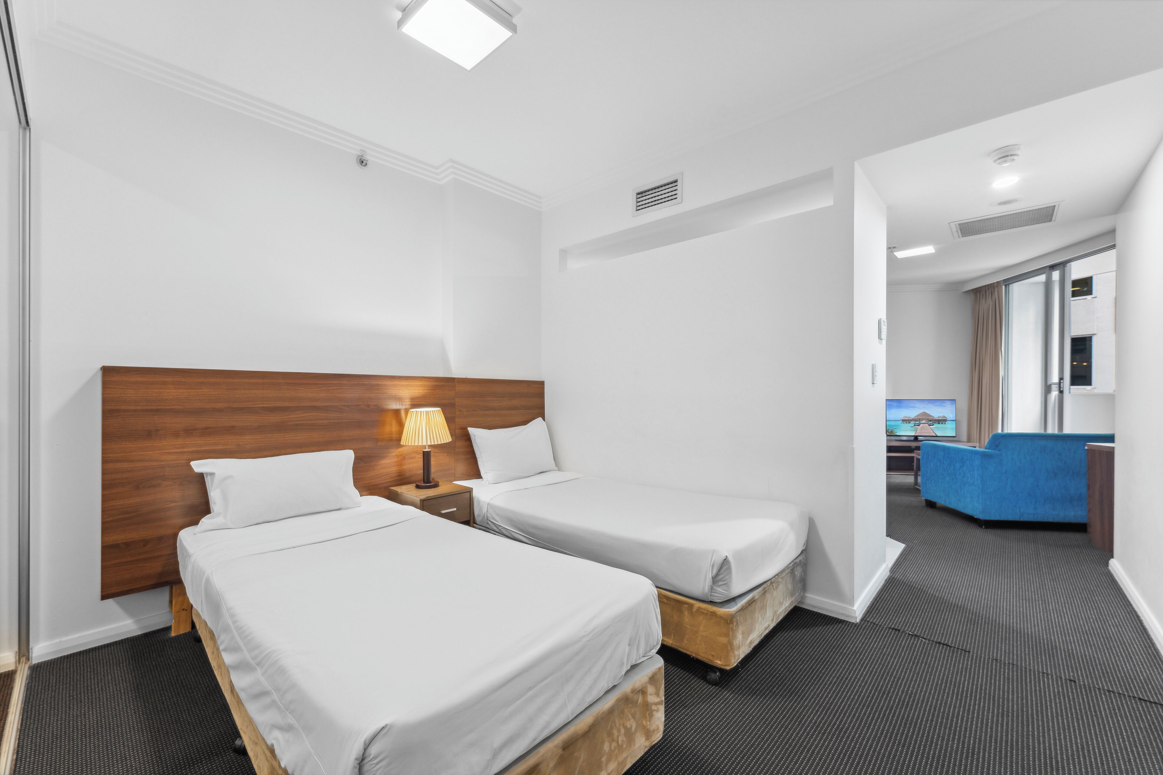 Une chambre d’hôtel avec deux lits simples, une tête de lit en bois, une table de chevet avec une lampe, et une vue sur un canapé bleu situé dans la chambre adjacente.
