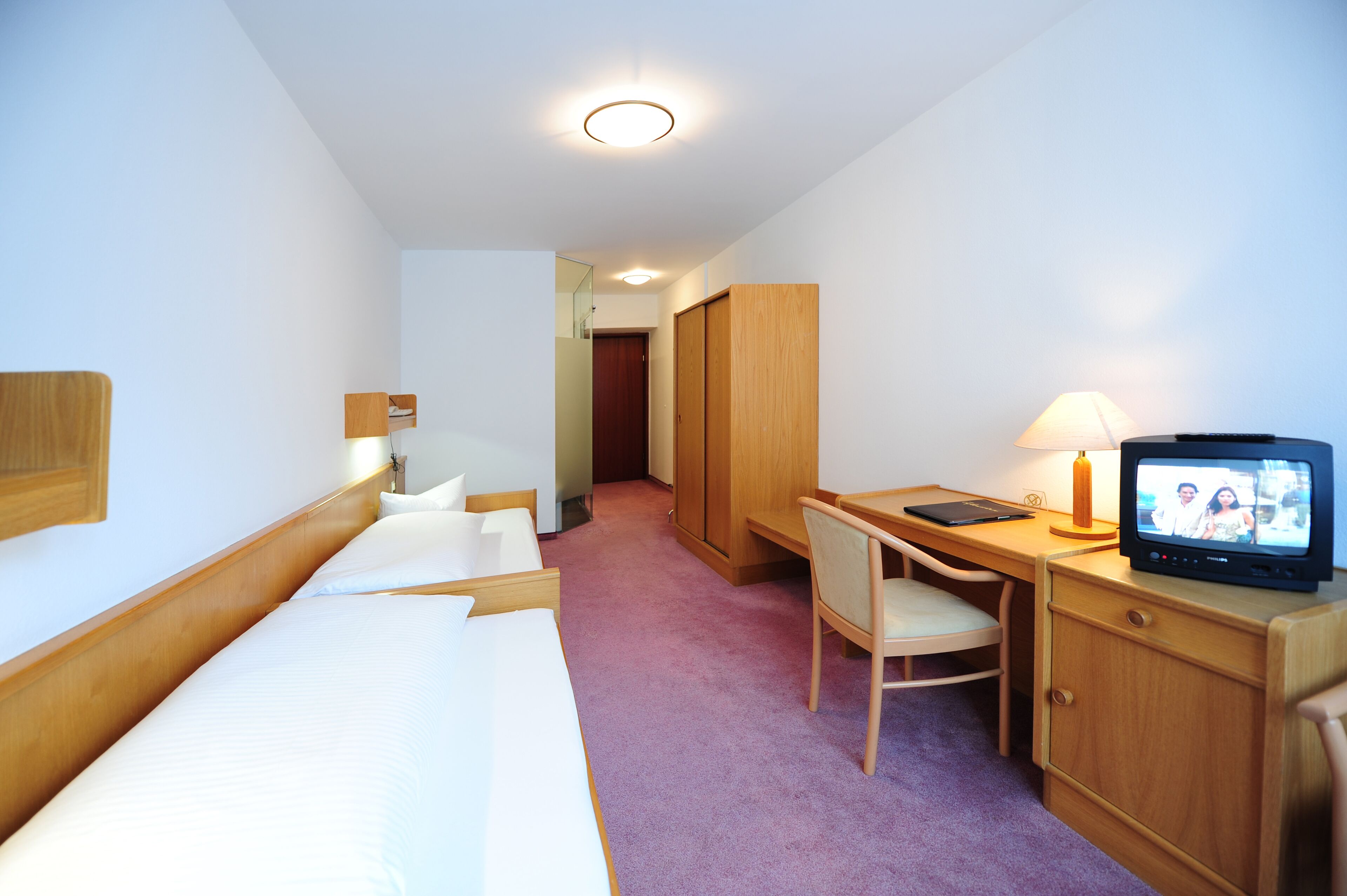 A hotel room with a bed, a desk with a chair, a television, and a wardrobe.