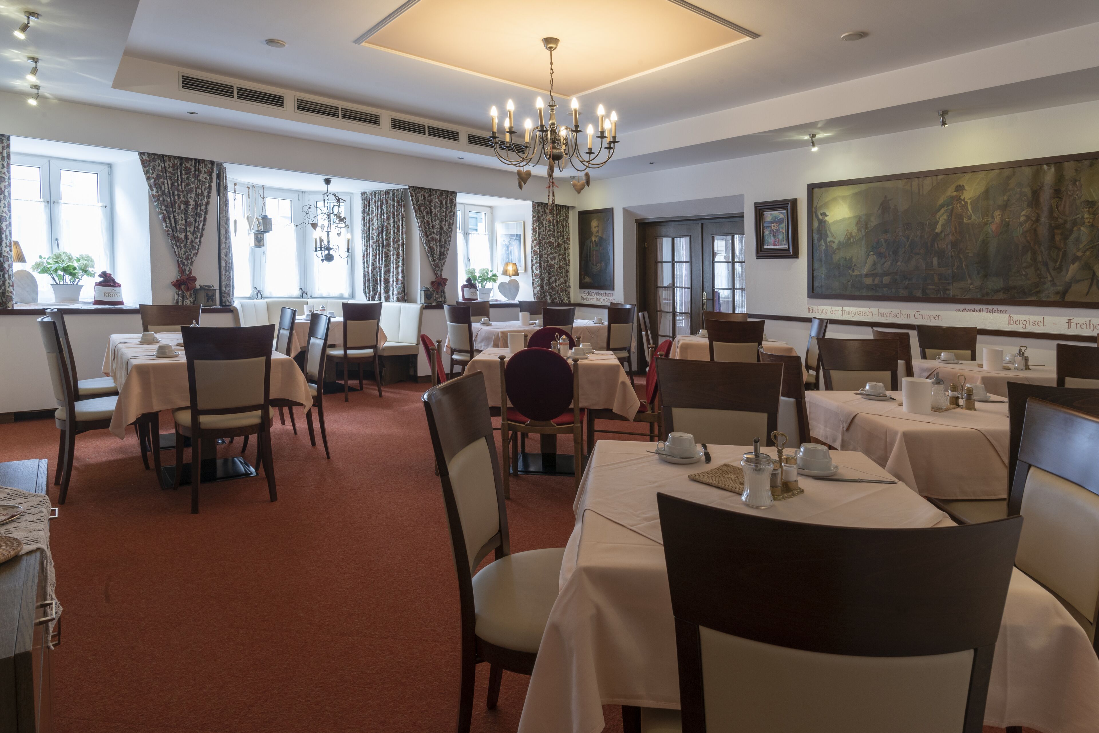 Une salle à manger avec des tables dressées, un lustre et un grand tableau accroché au mur.