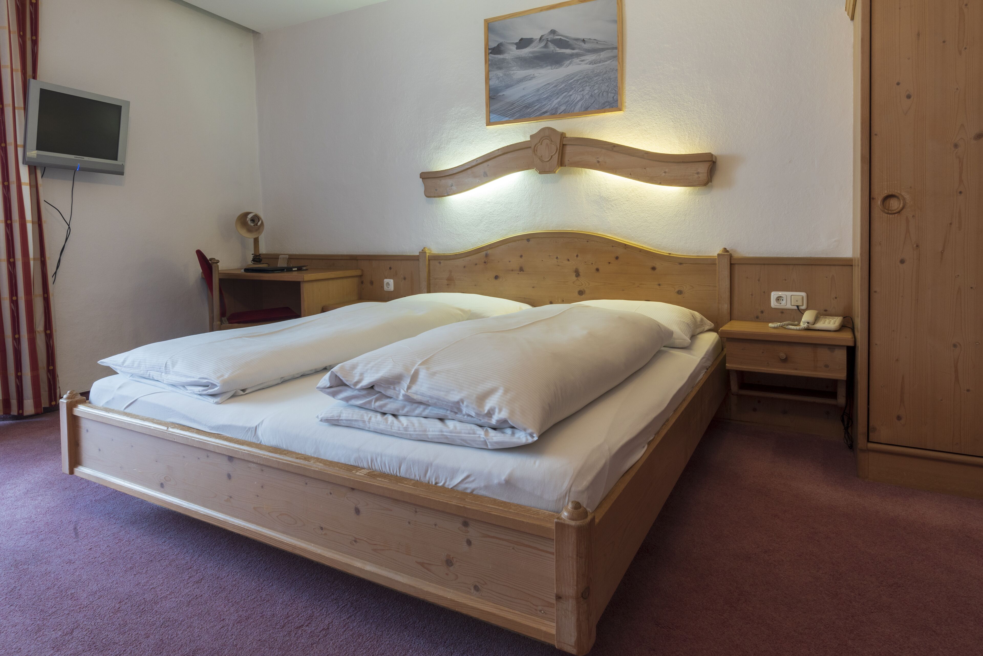 A wooden bed with two pillows, a bedside table with a lamp, a wall-mounted TV, and a painting above the bed.