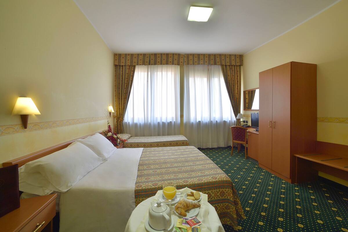 Habitación de hotel con dos camas, un escritorio, una silla, un armario y una bandeja de desayuno con cruasanes y jugo de naranja.