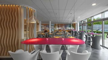 Ein modernes Restaurant mit einem zentralen rotem Tisch, umgeben von weißen Stühlen, einer gebogenen Holzwand und Pendelleuchten.