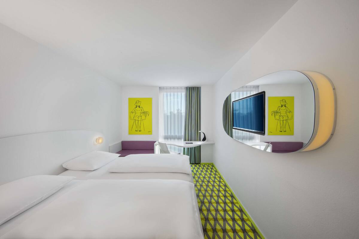 Une chambre d’hôtel équipée d’un lit, d’un bureau, d’une chaise, d’un miroir et d’œuvres d’art accrochées aux murs.