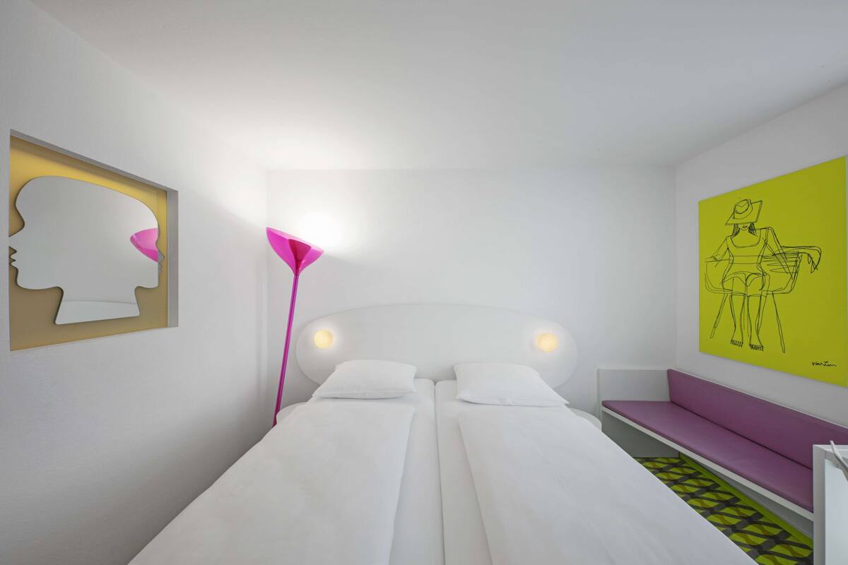 Une chambre à coucher moderne avec un lit, des lampes de chevet, un banc et des œuvres d’art accrochées aux murs.
