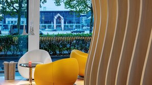 Ein modernes Interieur mit einer Sitzecke, bestehend aus einem weißen und zwei gelben Sesseln, einem kleinen Tisch mit Kerze sowie Blick auf ein Gebäude und die Straße.