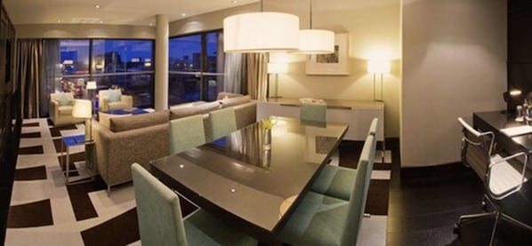 A modern hotel room with a dining area, a sofa, and a view of the cityscape at night.