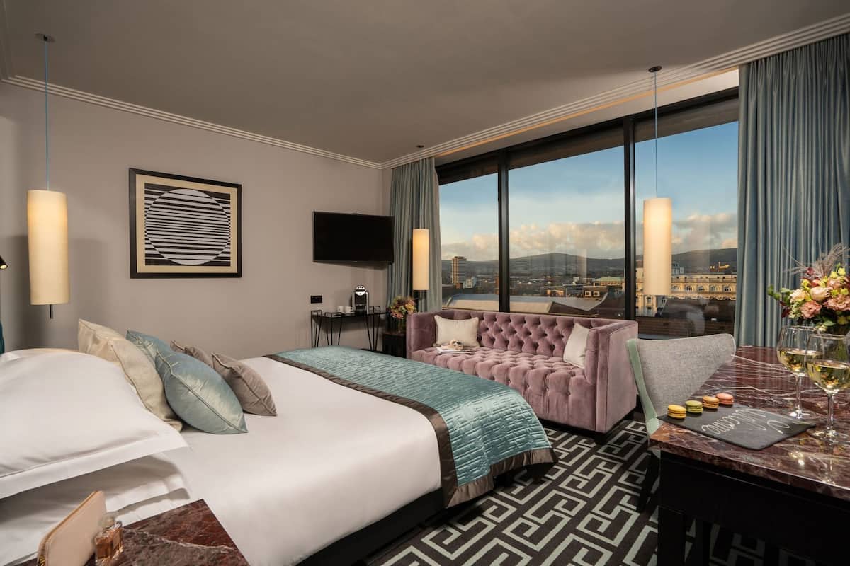 A hotel room with a bed, a sofa, a desk with a view, a TV, and a large window.