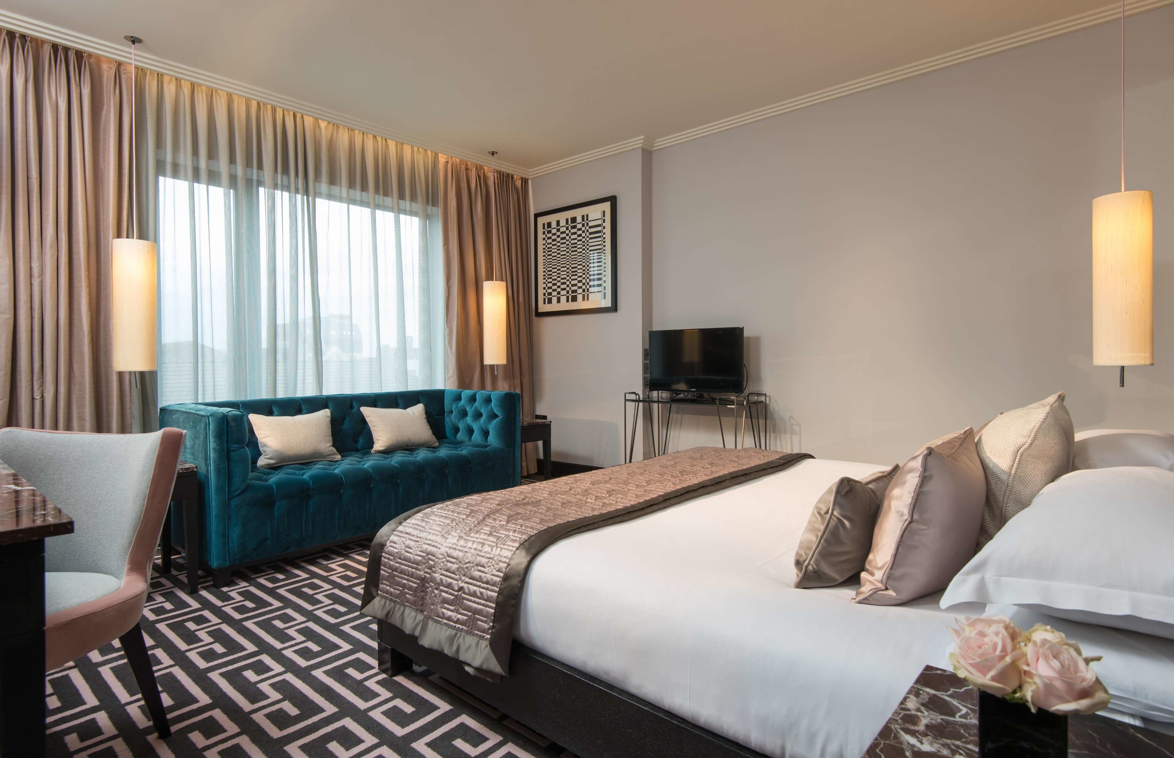 A hotel room with a large bed, a blue sofa, a desk with a chair, a TV, and a large window with curtains.