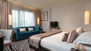 A hotel room with a large bed, a blue sofa, a desk with a chair, a TV, and a large window with curtains.