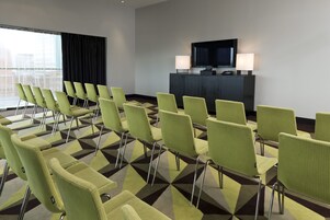 Un salón de conferencias con filas de sillas verdes, un mueble negro con un televisor y una alfombra estampada.