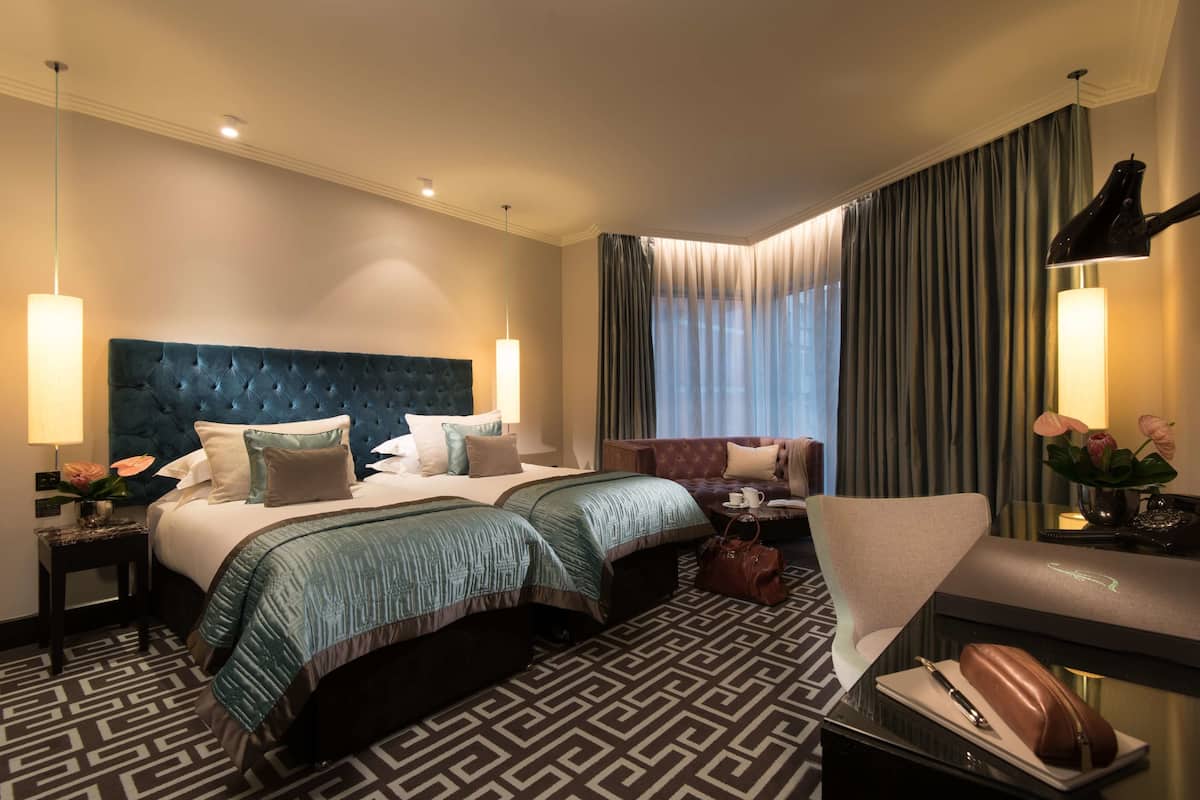 A hotel room with a large bed, two smaller beds, a sofa, a desk, and a chair. There are lamps, a vase with flowers, and a handbag on the desk.