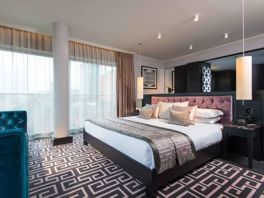 A modern hotel room with a large bed, a tufted headboard, a patterned floor, and a large window with curtains.