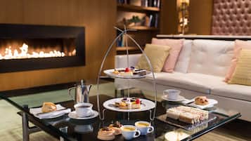 A glass-top table with a three-tiered dessert stand, a coffee pot, and various pastries and sandwiches.