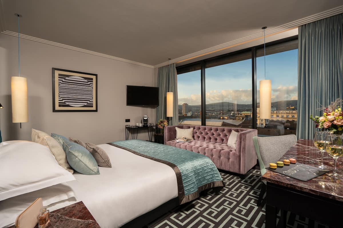 A hotel room with a large bed, a sofa, a desk with a laptop, and a view of the city.