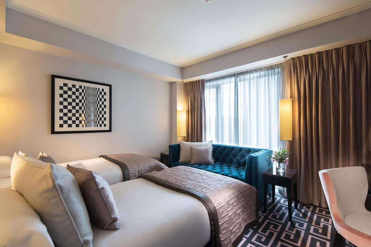 A hotel room with a checkered wall art, a blue upholstered sofa, a bed with multiple pillows, and a chair.