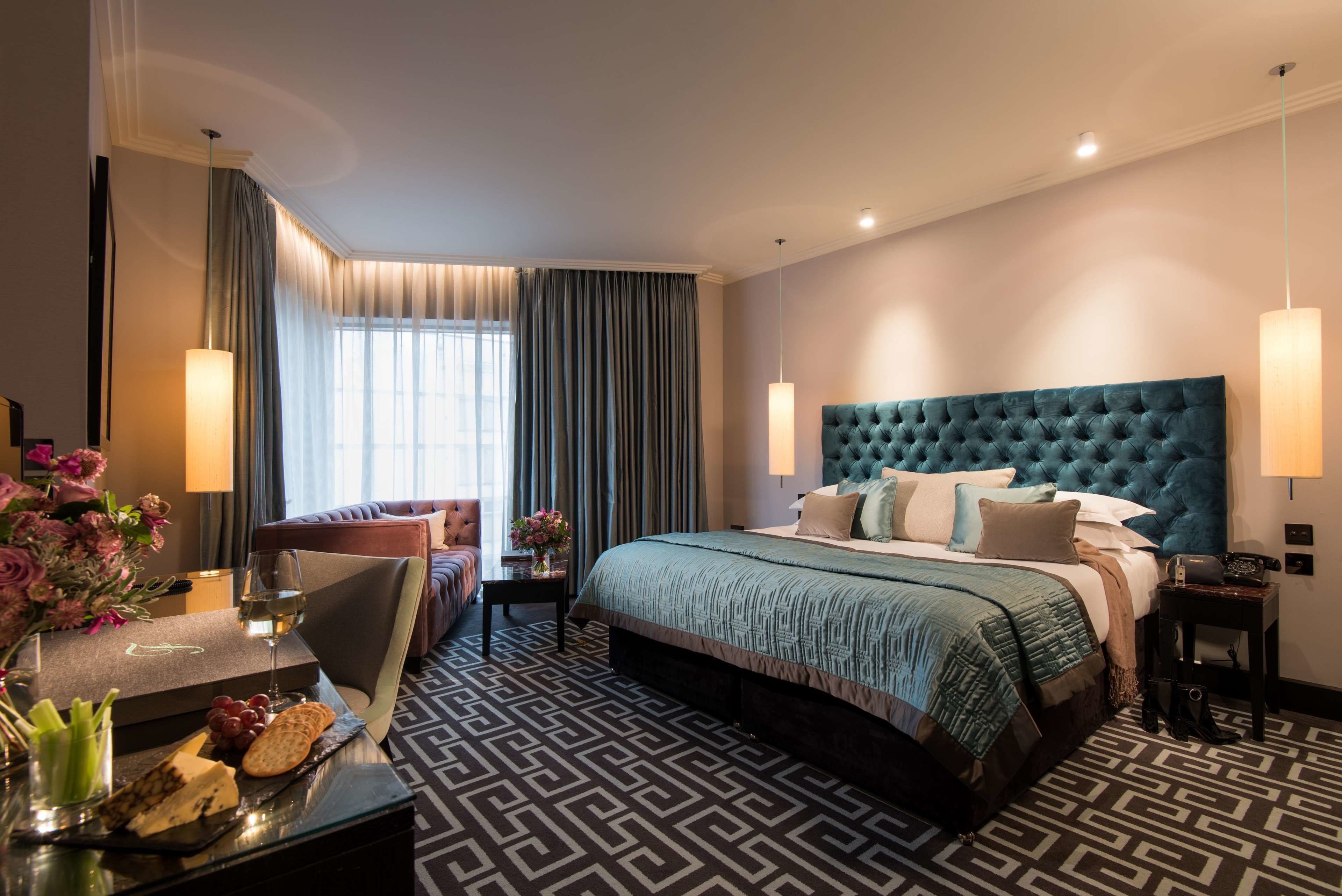 Een hotelkamer met een groot bed, een zithoek met een fauteuil, een bureau met een glas wijn en een bloevas.