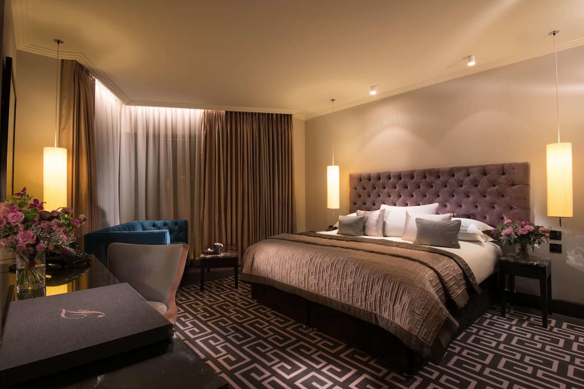 A hotel room with a large bed, a tufted headboard, a patterned floor, a desk with a chair, and two bedside lamps.