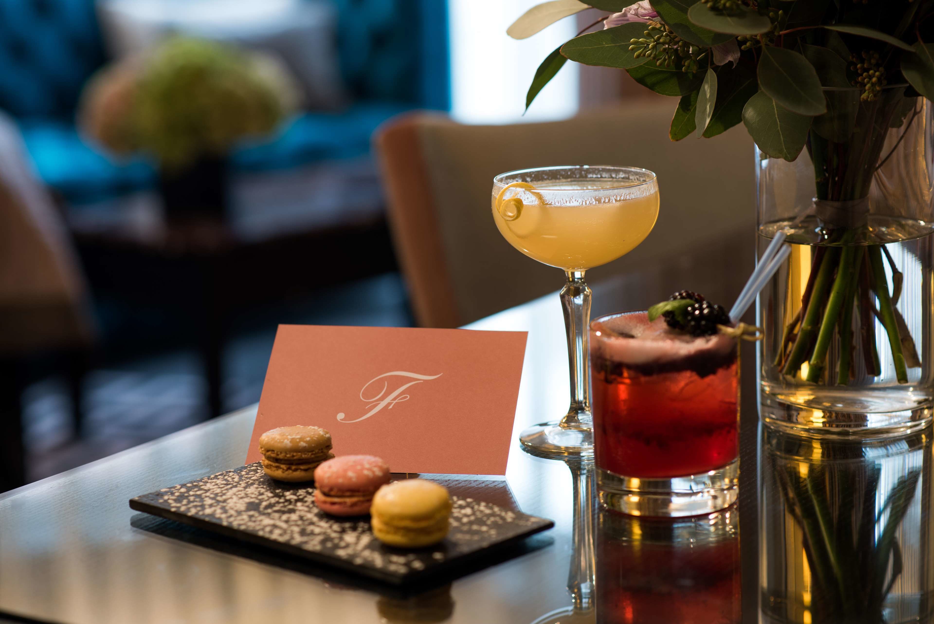 A table with a cocktail glass, a martini glass, a dessert plate with macarons, a card, and a vase with flowers.