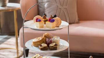 A three-tiered dessert stand with various pastries and sandwiches, a teapot, and a cup and saucer on a glass table.