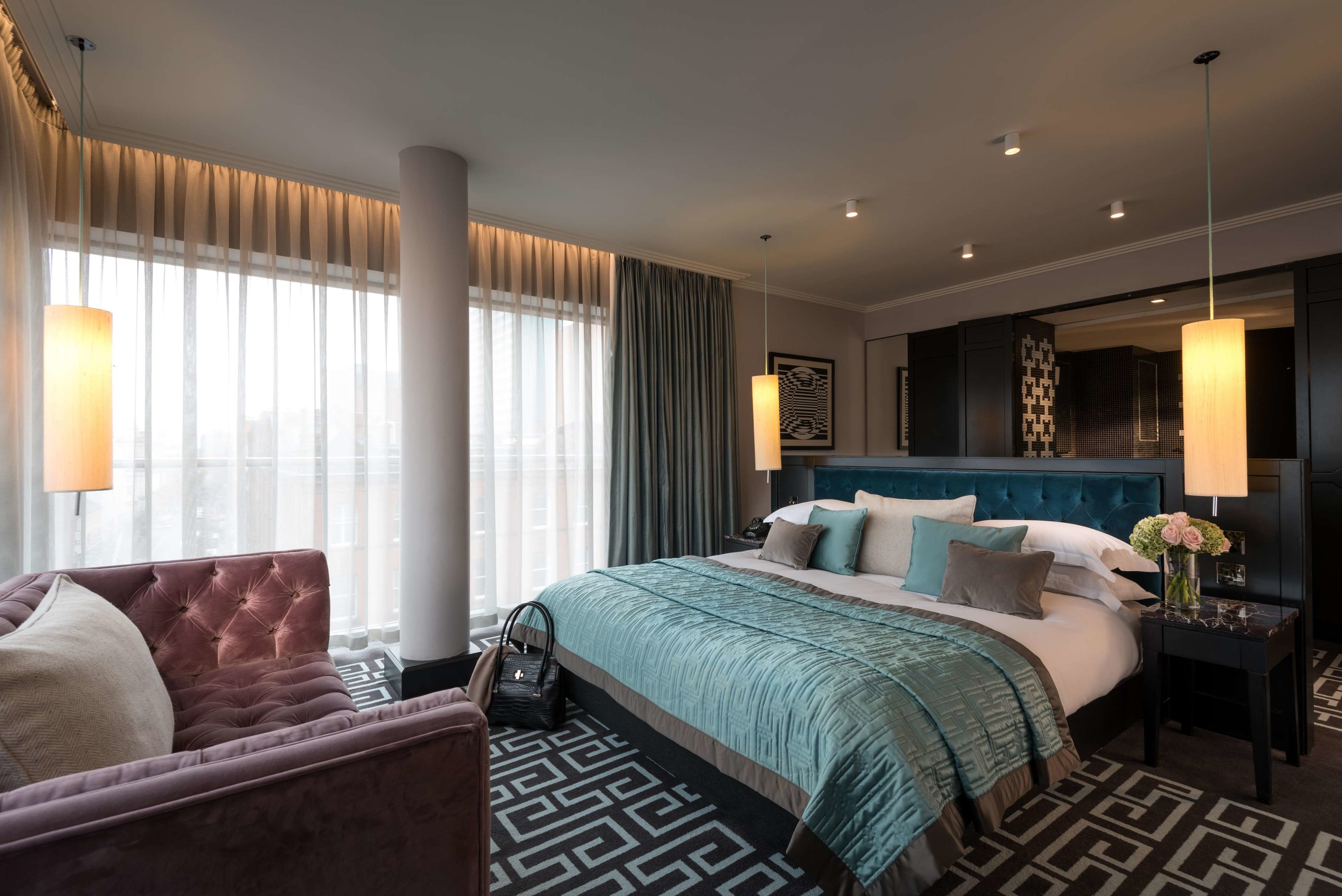 A modern hotel room with a large bed, a sitting area with a sofa, and a view of the city through large windows.
