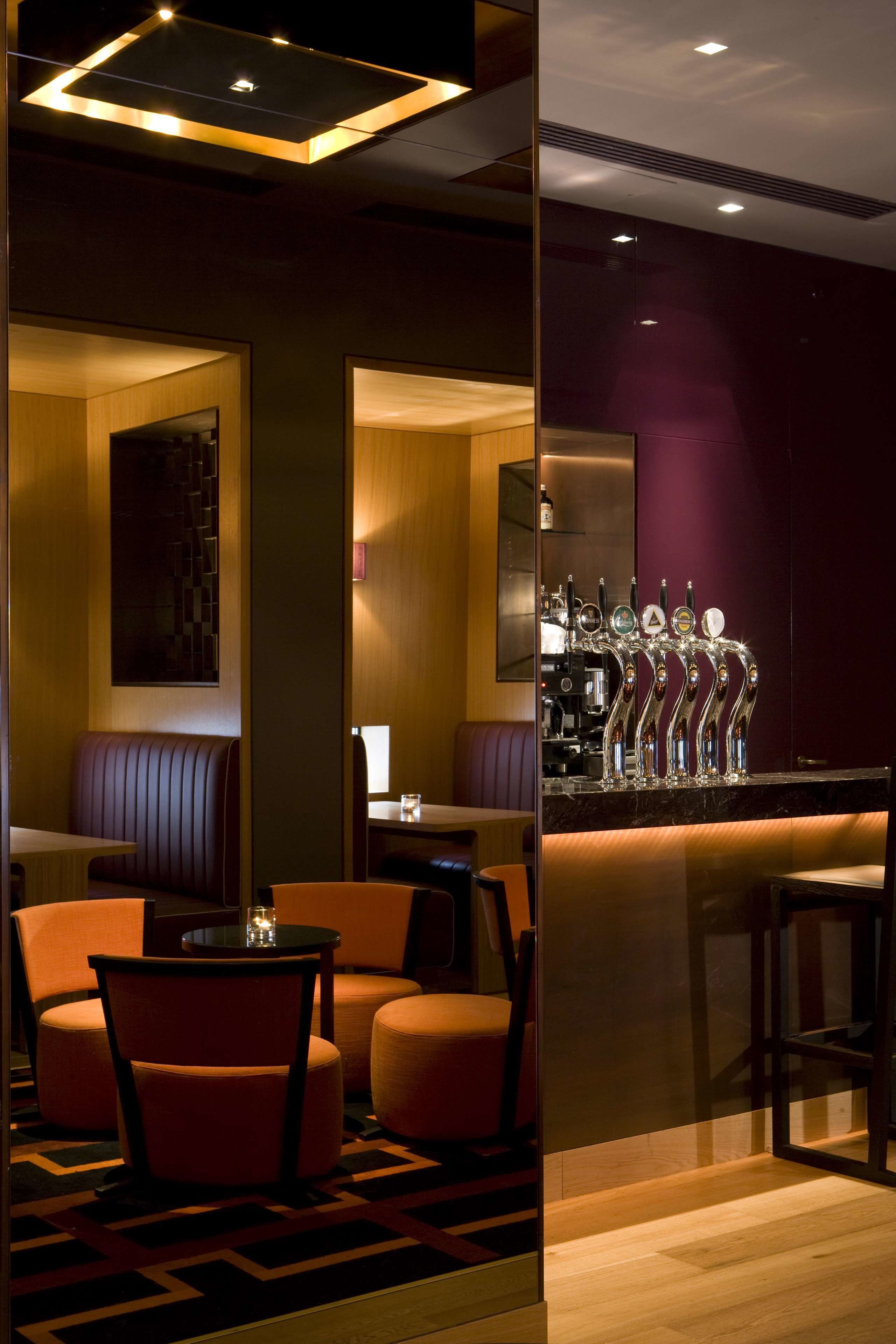 A modern bar area with a row of illuminated taps, seating with orange chairs, and a patterned floor.