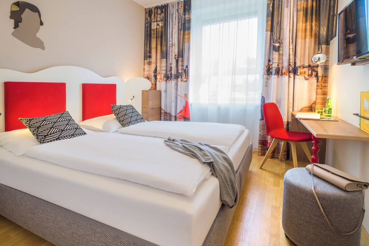 Une chambre d’hôtel avec deux lits, un bureau, une chaise, une valise et une grande fenêtre avec des rideaux.