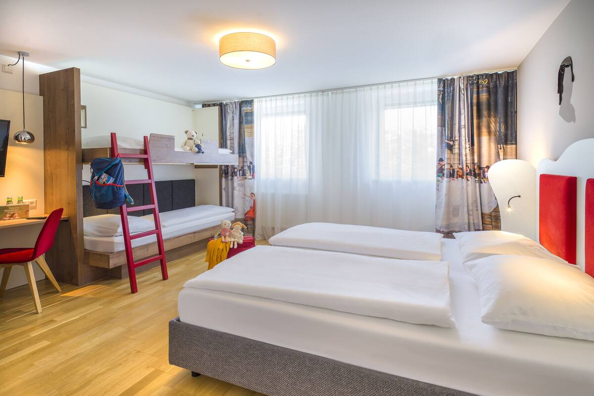 Une chambre d’hôtel avec un lit superposé, un bureau avec une chaise, une chaise rouge, une lampe et une grande fenêtre avec des rideaux.