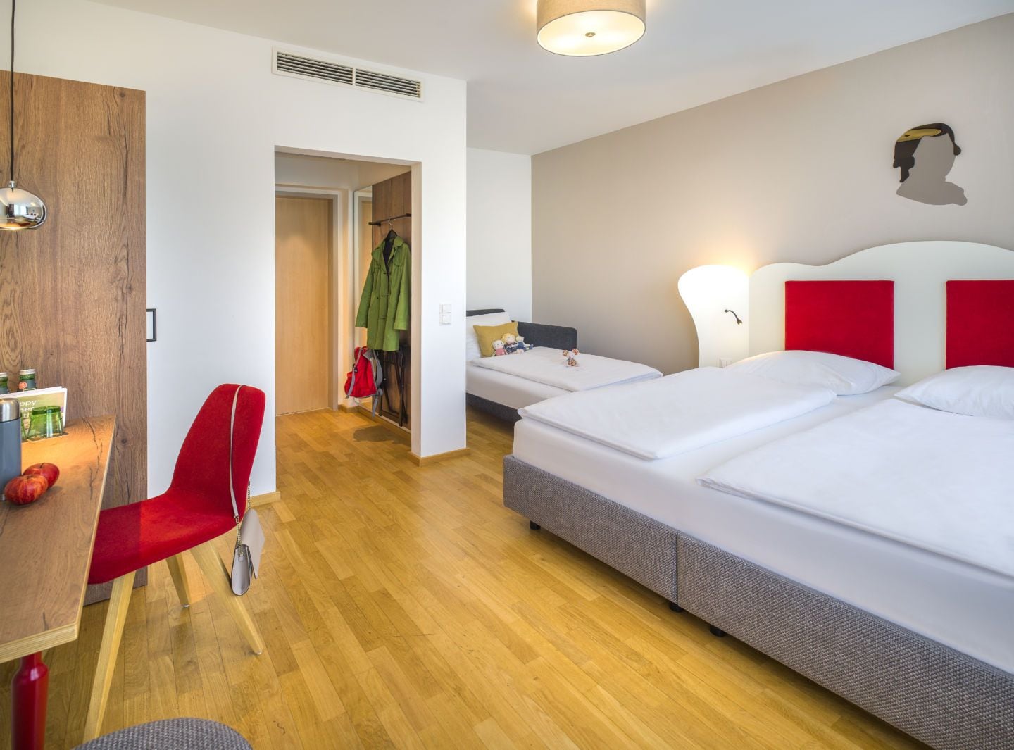 A hotel room with a wooden floor, a desk with a chair, two beds with red headboards, and a coat rack with clothes.