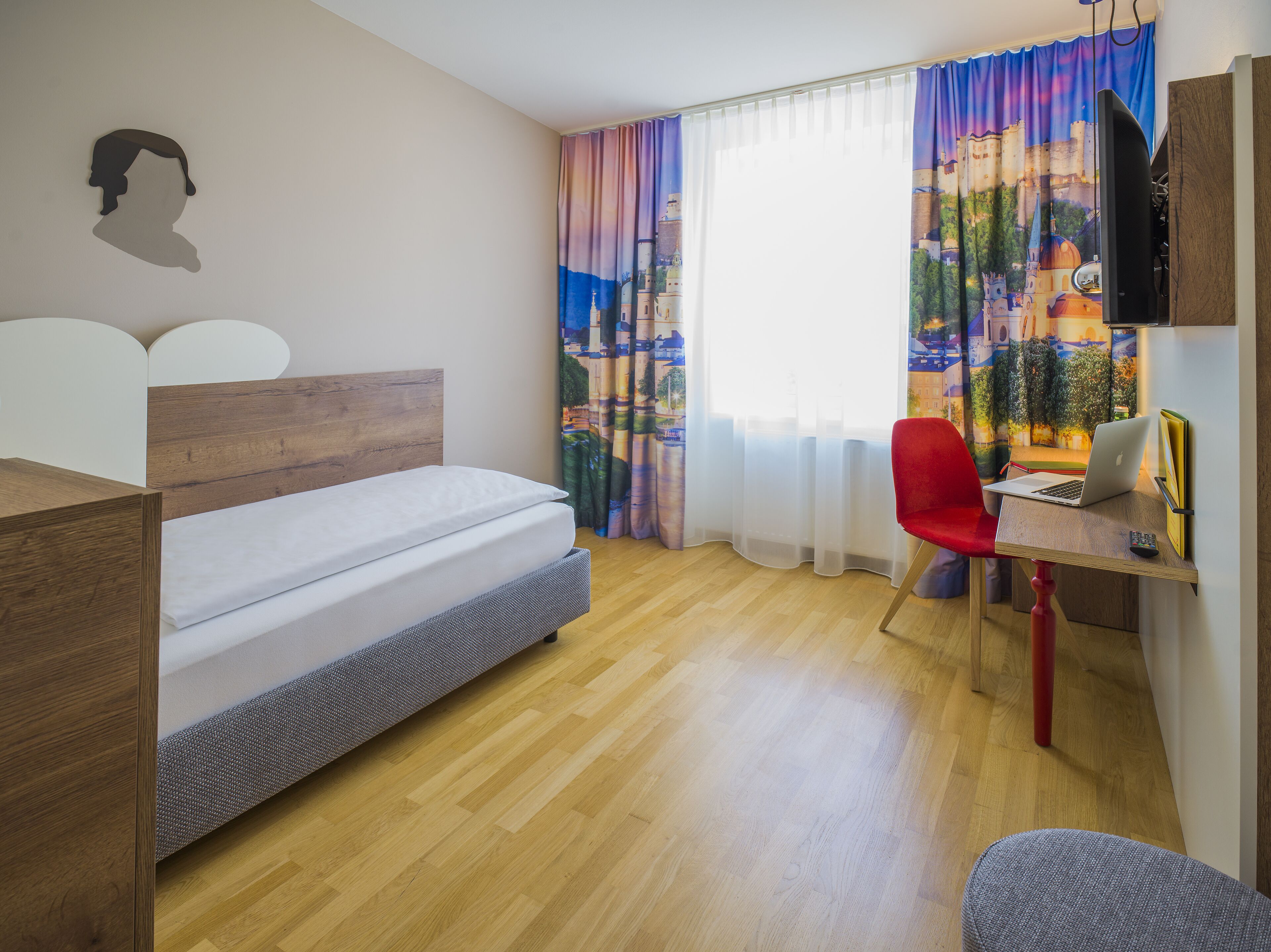 A bedroom with a bed, a desk with a laptop, a red chair, and a TV on a wooden stand.