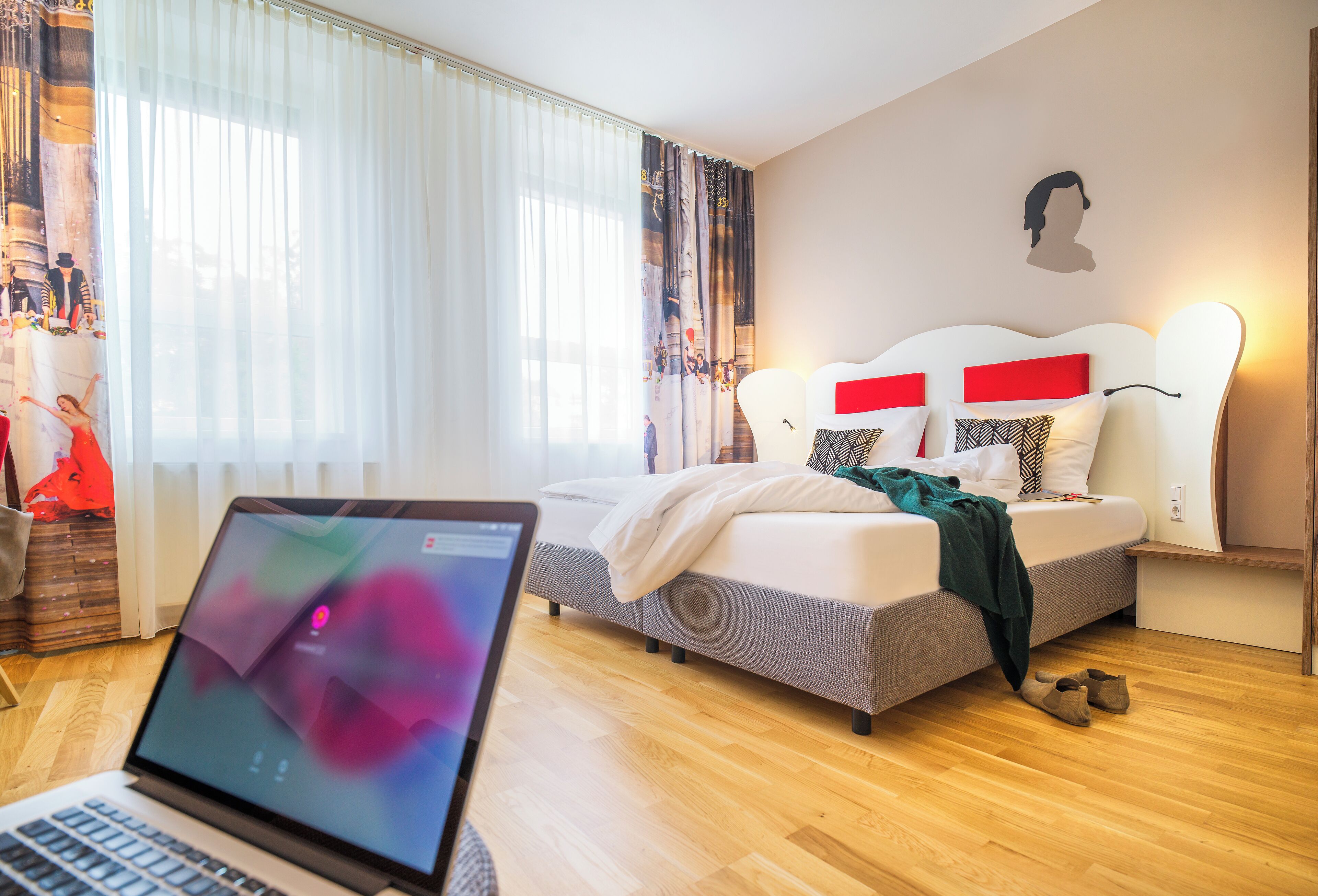 A modern bedroom with a bed, a laptop on a side table, and a large window with sheer curtains.