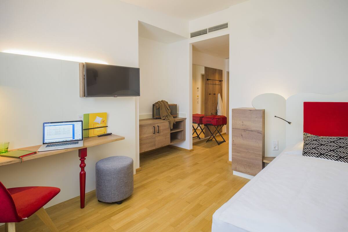 Une chambre d’hôtel moderne avec un plancher en bois, un bureau avec un ordinateur portable, un lit avec une tête de lit rouge et un téléviseur fixé au mur.