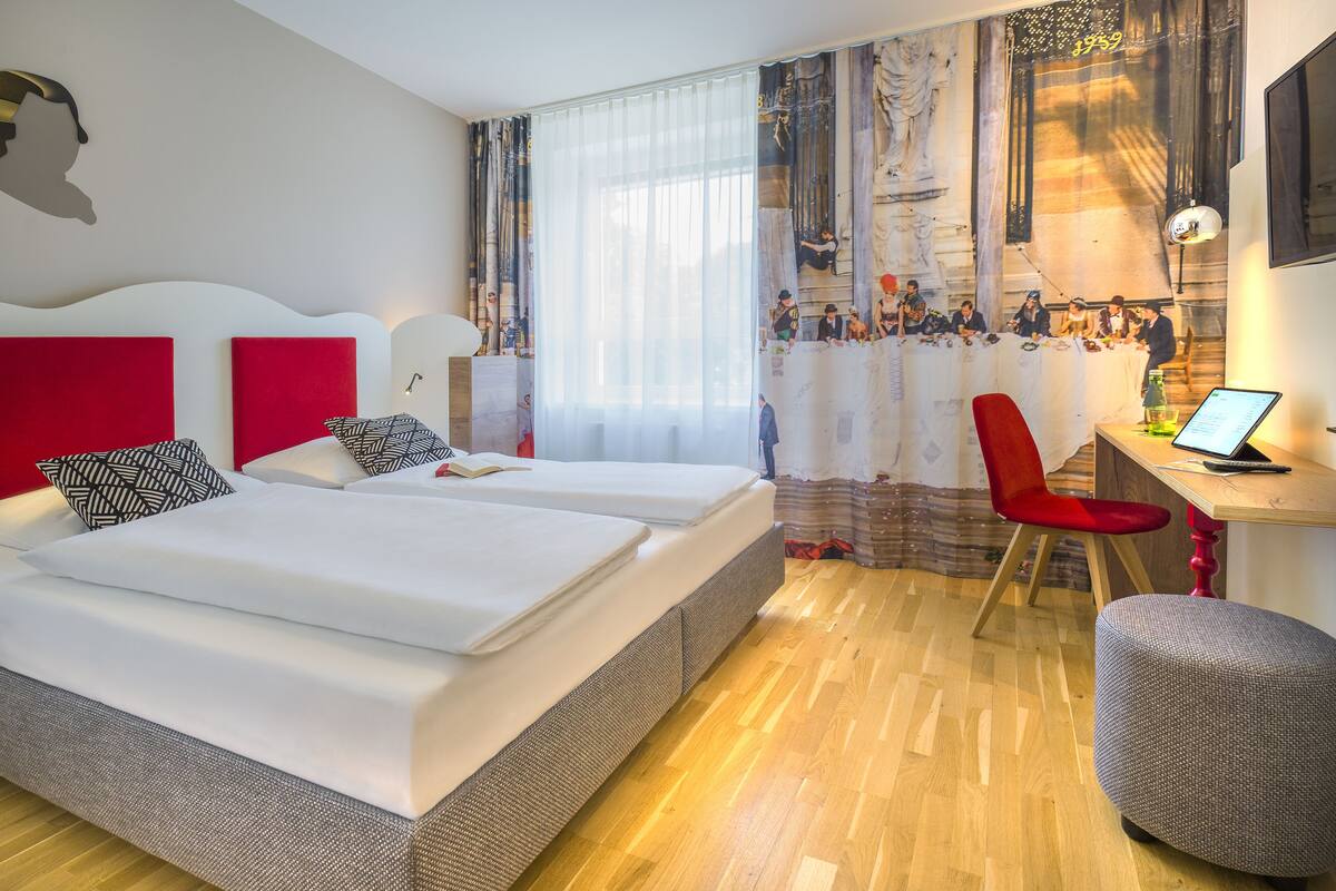 Une chambre d’hôtel avec deux lits, un bureau avec un ordinateur portable, une chaise rouge et un pouf rouge.