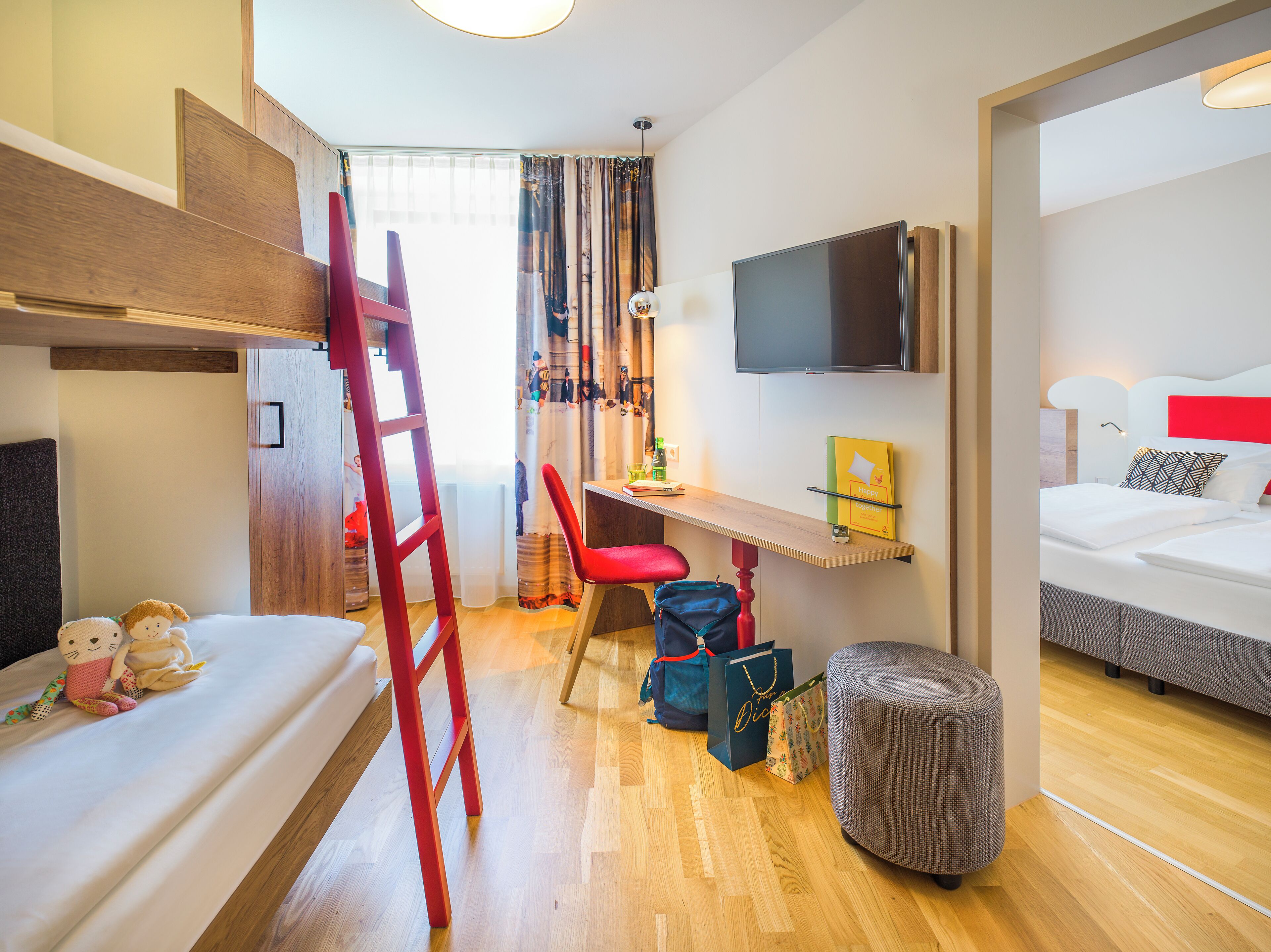 A hotel room with a bunk bed, a desk with a chair, a TV mounted on the wall, and a small ottoman.