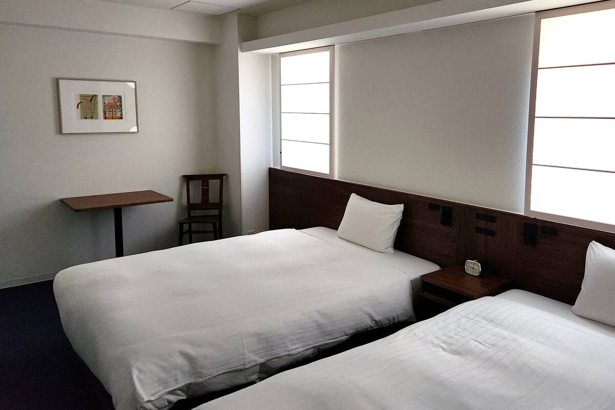 Une chambre d’hôtel avec deux lits, une table de chevet en bois, une petite table et une fenêtre avec des stores.