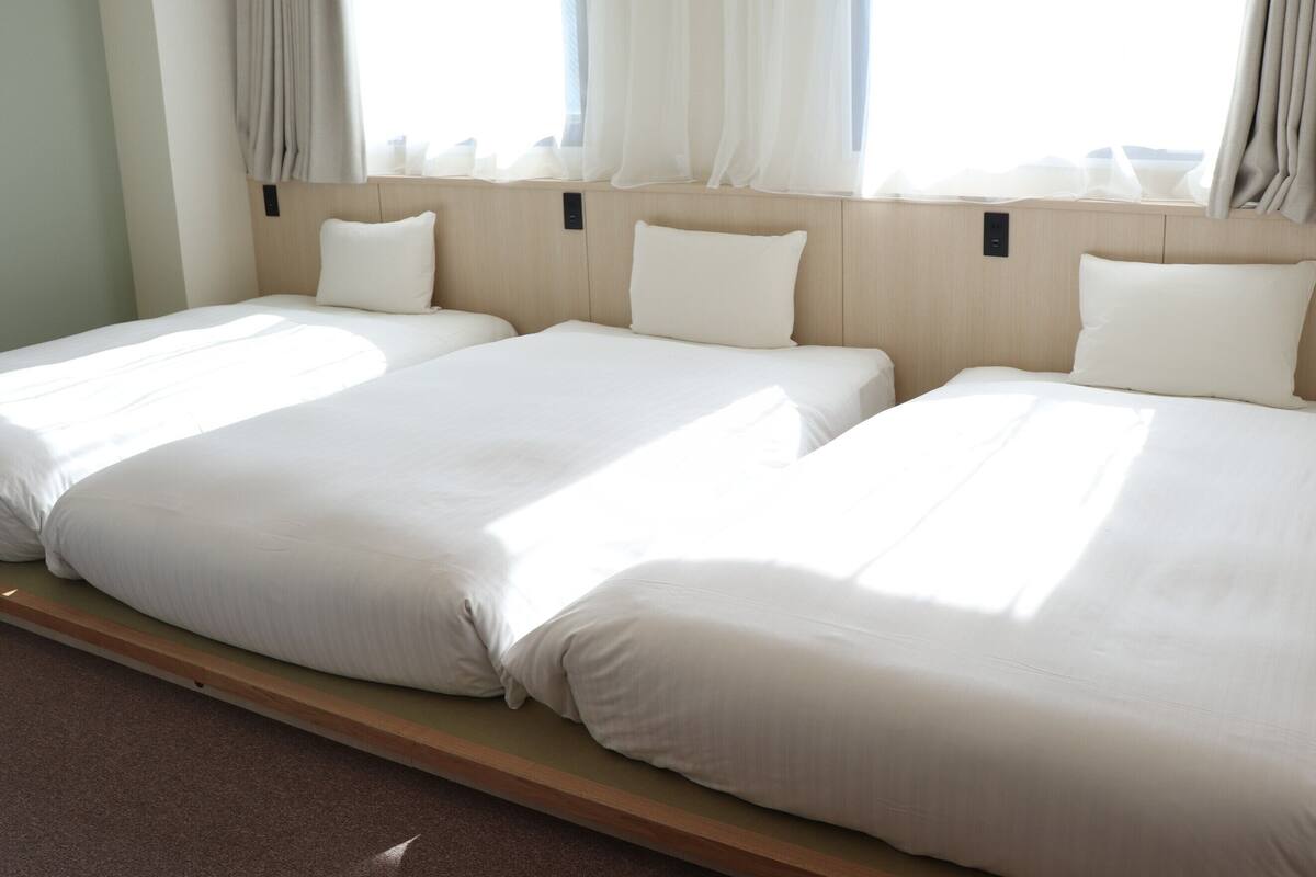 Une chambre d’hôtel avec deux lits simples, une tête de lit en bois et une fenêtre ornée de rideaux légers.