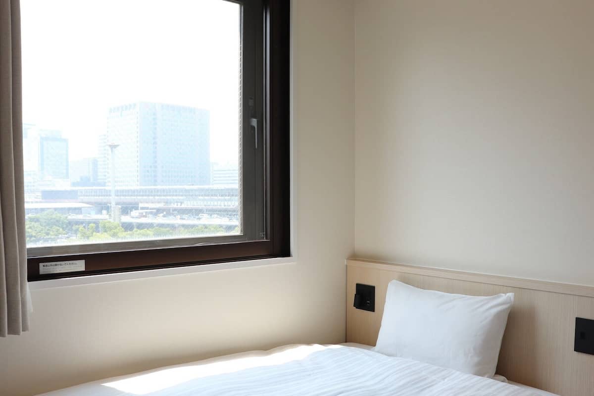 Une chambre d’hôtel avec un lit, une fenêtre donnant sur des immeubles et un interrupteur fixé au mur.