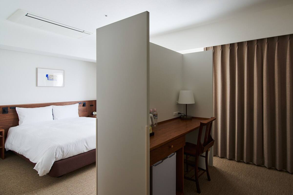 Une chambre d’hôtel avec un lit, un bureau, une chaise, une lampe et une fenêtre avec des rideaux.