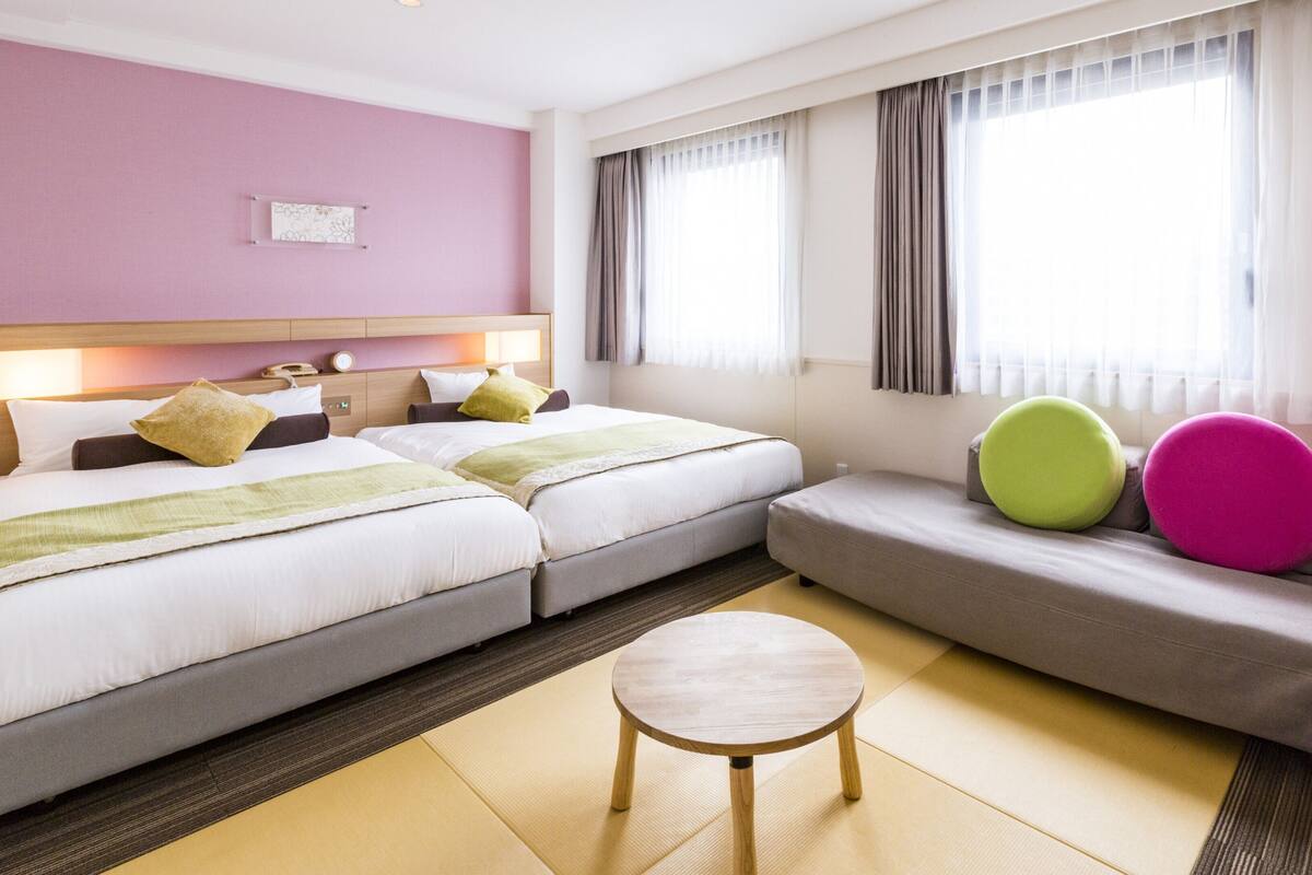 Une chambre d’hôtel avec deux lits, un canapé, un petit tabouret en bois et une fenêtre avec des rideaux.