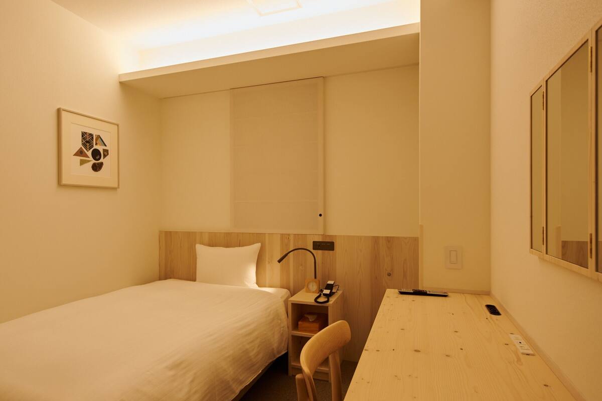 Une petite chambre d’hôtel, bien rangée, avec un lit simple, un bureau, une chaise et une table de chevet sur laquelle se trouvent une lampe et une horloge.