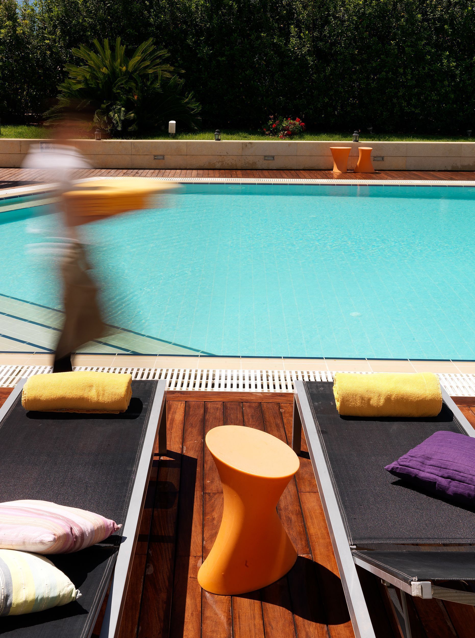 A poolside area with lounge chairs and a small orange table, surrounded by greenery and a tiled deck.