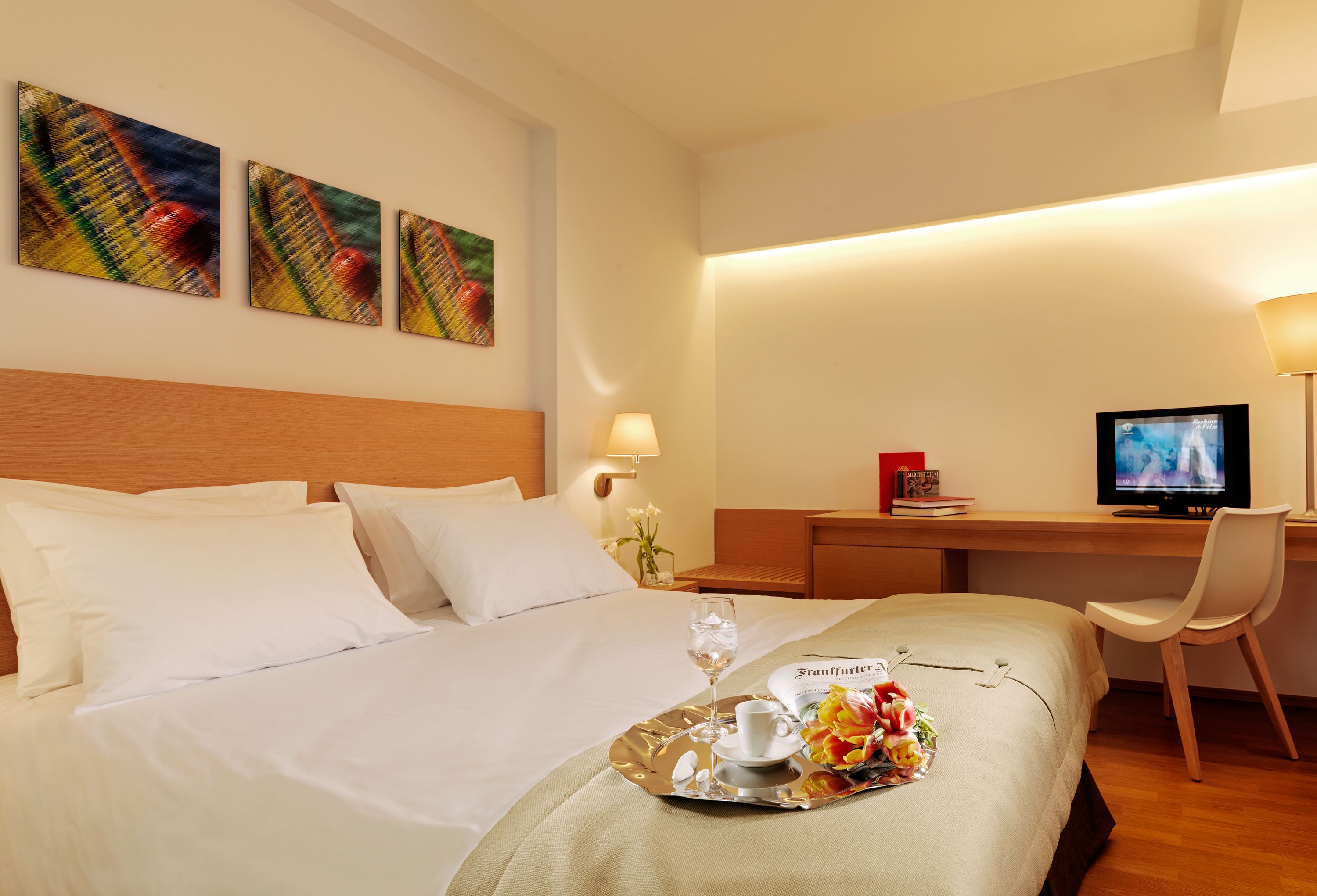 A hotel room with a bed, a desk with a computer, a TV, and a tray with a drink and flowers.