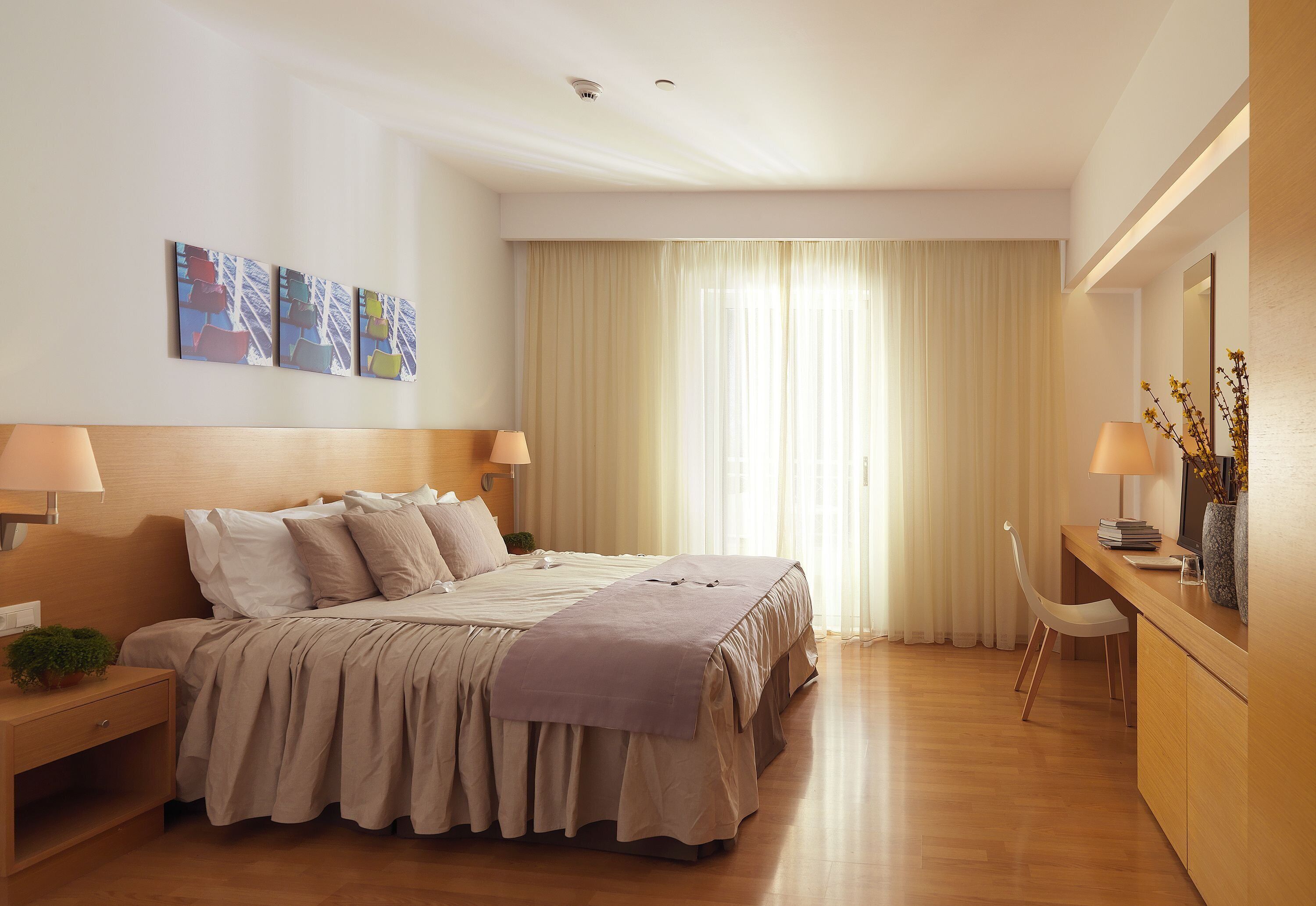 A modern hotel room with a large bed, bedside lamps, a desk with a chair, and a window with curtains.