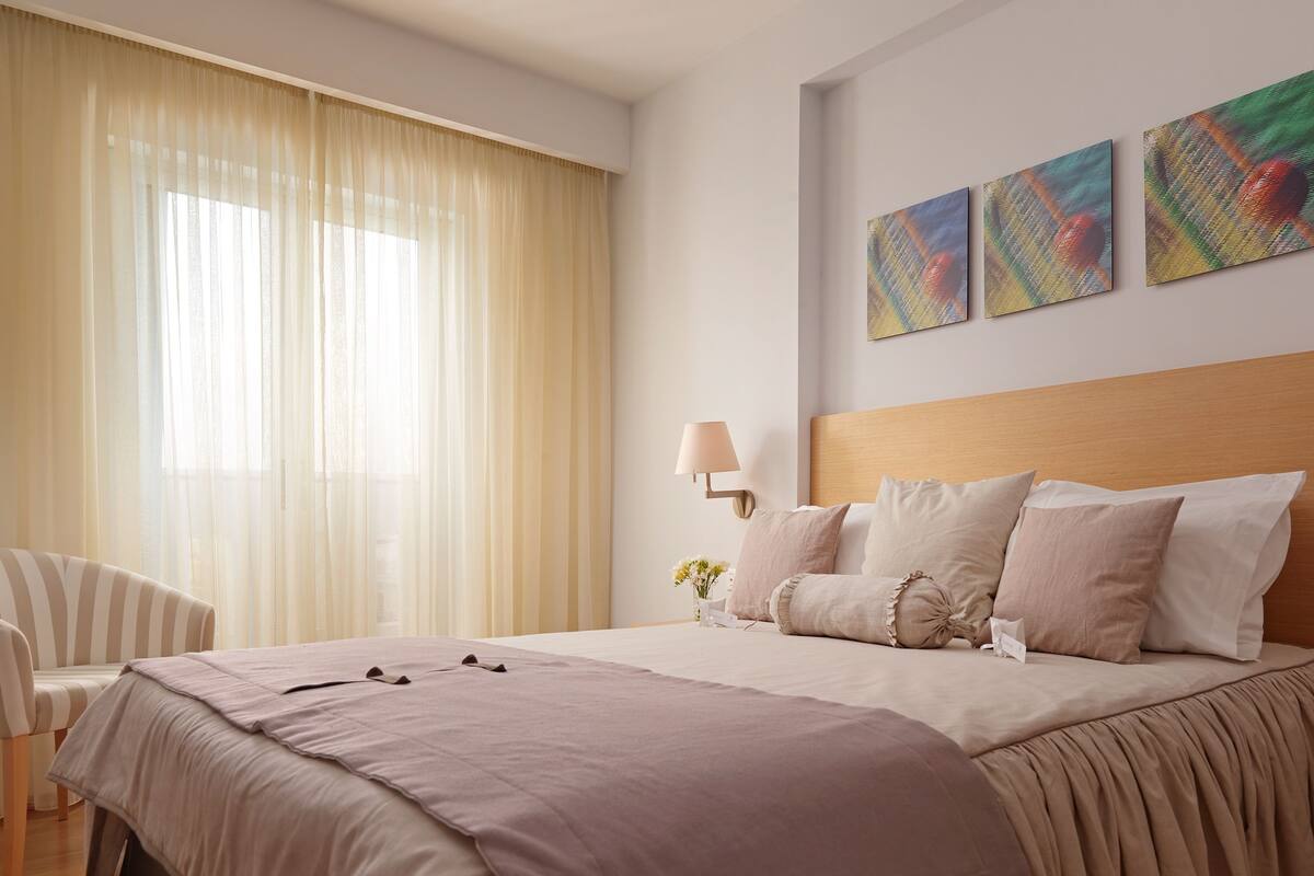 A hotel room with a large bed, a bedside table, a lamp, and a window with sheer curtains.