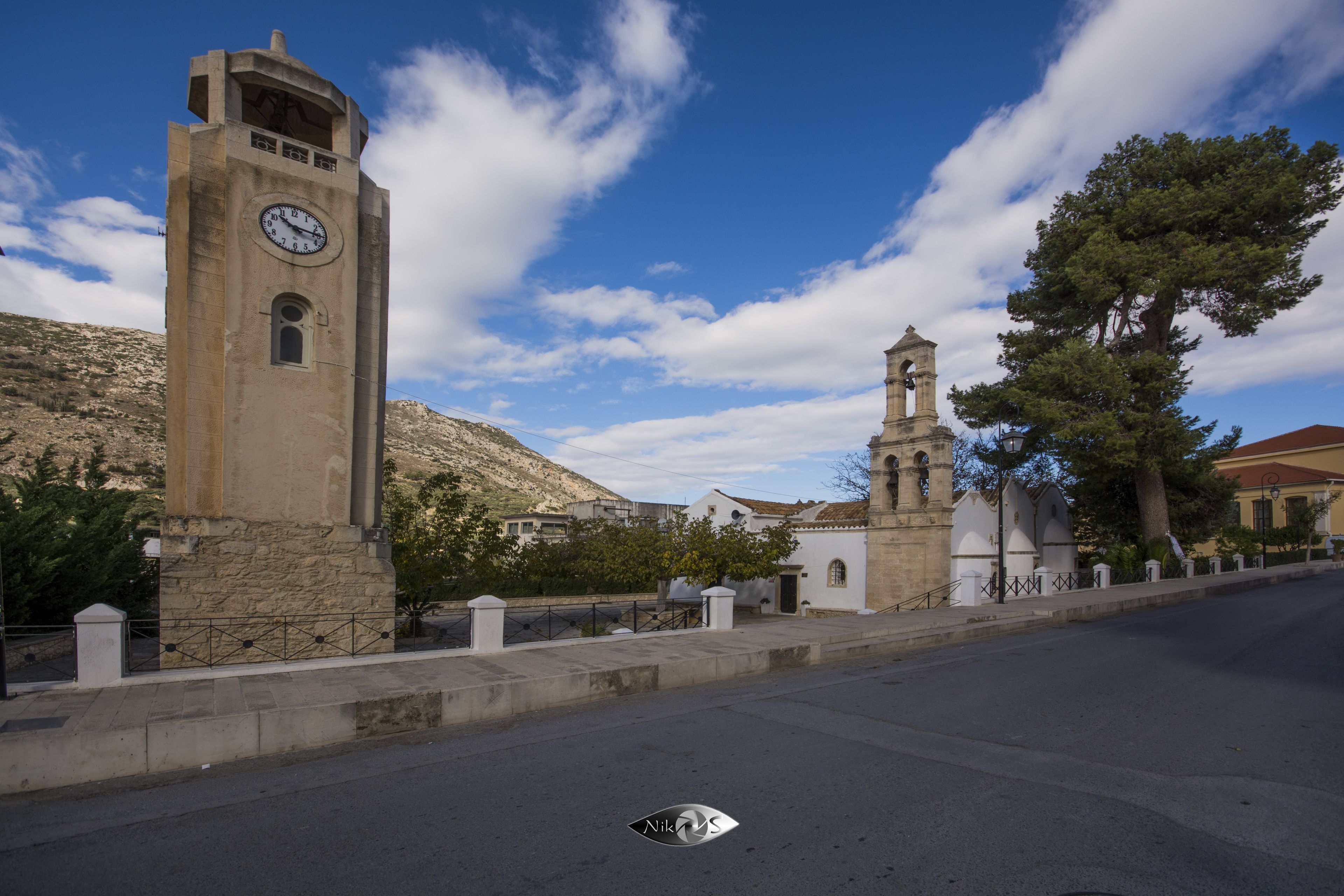 A street with a clock tower and a bell tower, surrounded by buildings and trees.