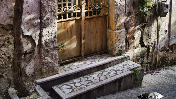 Un bâtiment en pierre avec un portail en fer forgé, des marches menant à une porte et un arbre aux fleurs violettes.