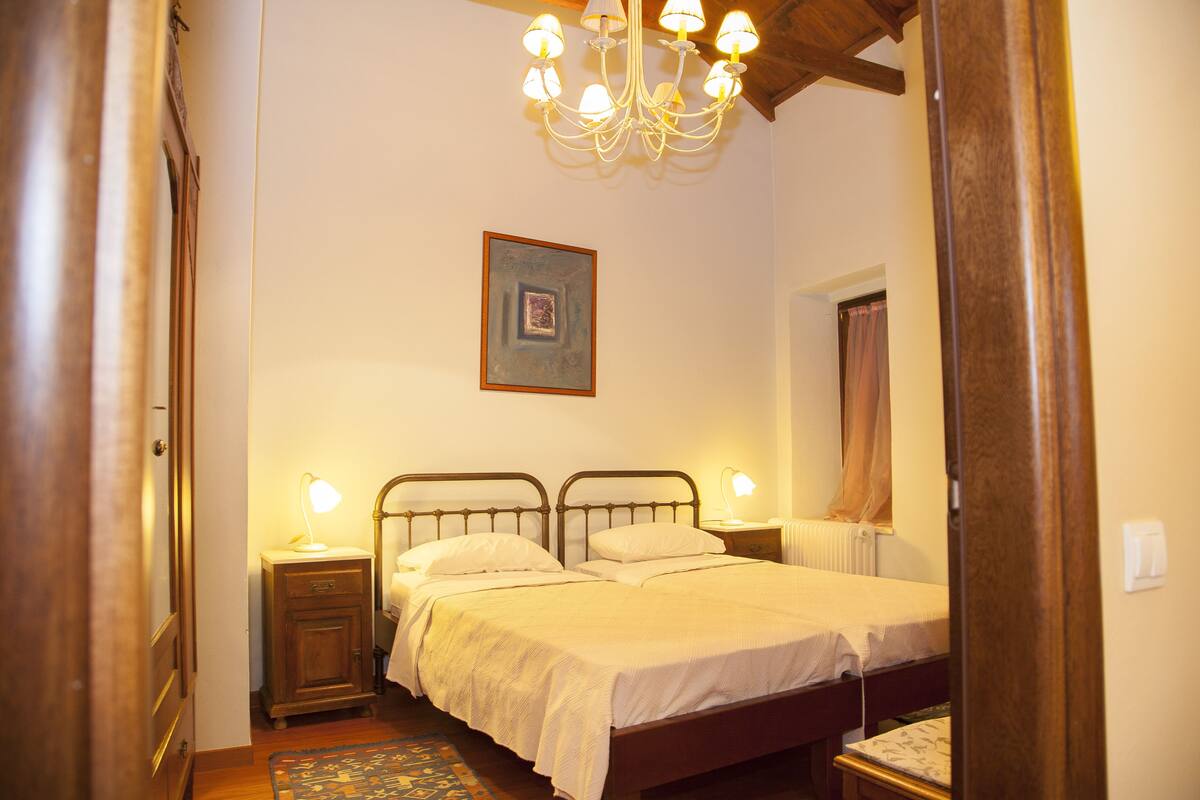 Ein Schlafzimmer mit einem Doppelbett, zwei Nachttischen mit Lampen, einer Kristallleuchtre, einem gerahmten Bild an der Wand und einer Holztür.