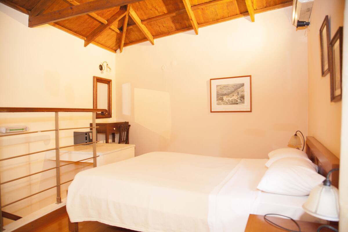 Ein Schlafzimmer mit Holzbalkendecke, einem Bett mit weißer Bettwäsche, einem Nachttisch mit Lampe, einem gerahmten Bild an der Wand und einem Spiegel über der Kommode.