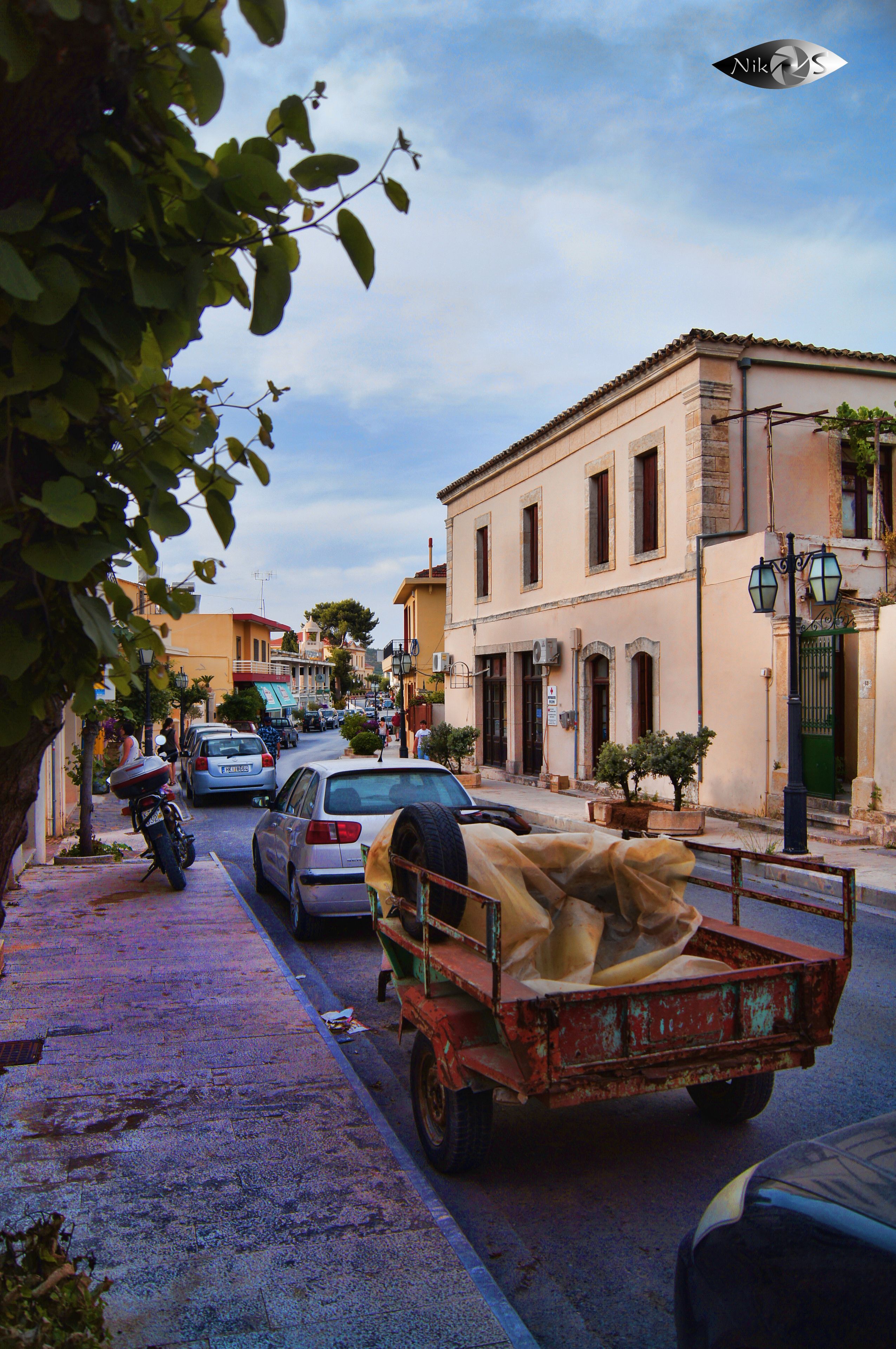 A street scene with parked cars, a motorcycle, and a cart with a large piece of cloth. Buildings line the street, and there are trees and plants along the sidewalk.