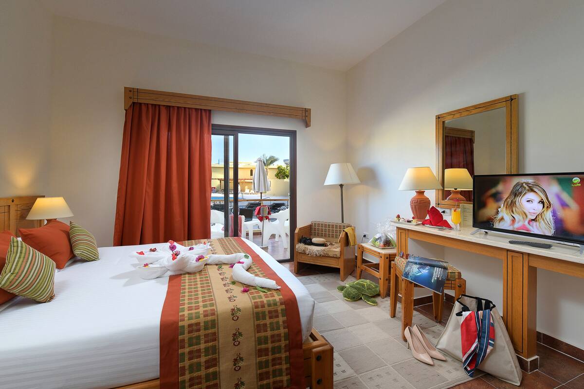 Habitación de hotel con cama, escritorio con televisor, silla y balcón con vistas.