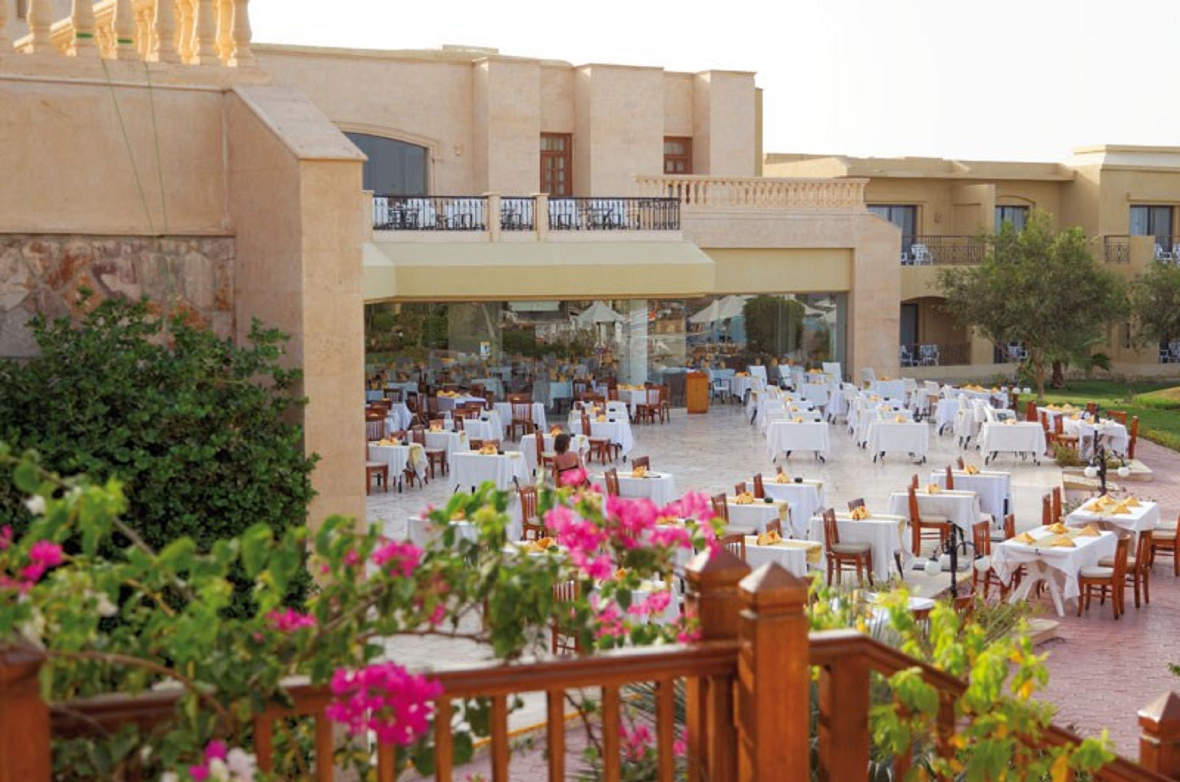 An outdoor dining area with multiple tables and chairs, surrounded by greenery and a building with balconies.